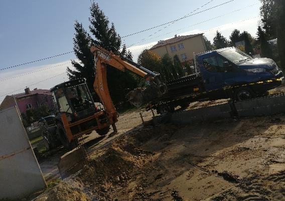 Pomarańczowa koparko-ładowarka załadowywana na lawetę transportową na placu budowy, w tle zabudowa mieszkalna i drzewa.