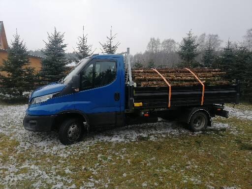 Niebieski samochód dostawczy z czarną skrzynią załadowany drewnem opałowym zabezpieczonym pomarańczowymi pasami transportowymi, stojący na trawie pokrytej śniegiem, w tle budynek i drzewa.