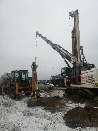 Ciężki sprzęt budowlany na placu budowy w zimowej scenerii: koparko-ładowarka, żuraw z ramieniem podnoszącym rurę oraz palownica HR 40, widoczne ślady opon na śniegu i wykopana ziemia.