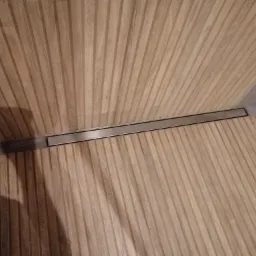 Ściana i podłoga wykończone panelami z drewna o pionowym wzorze, z widocznym liniowym odpływem prysznicowym w metalowej ramie.