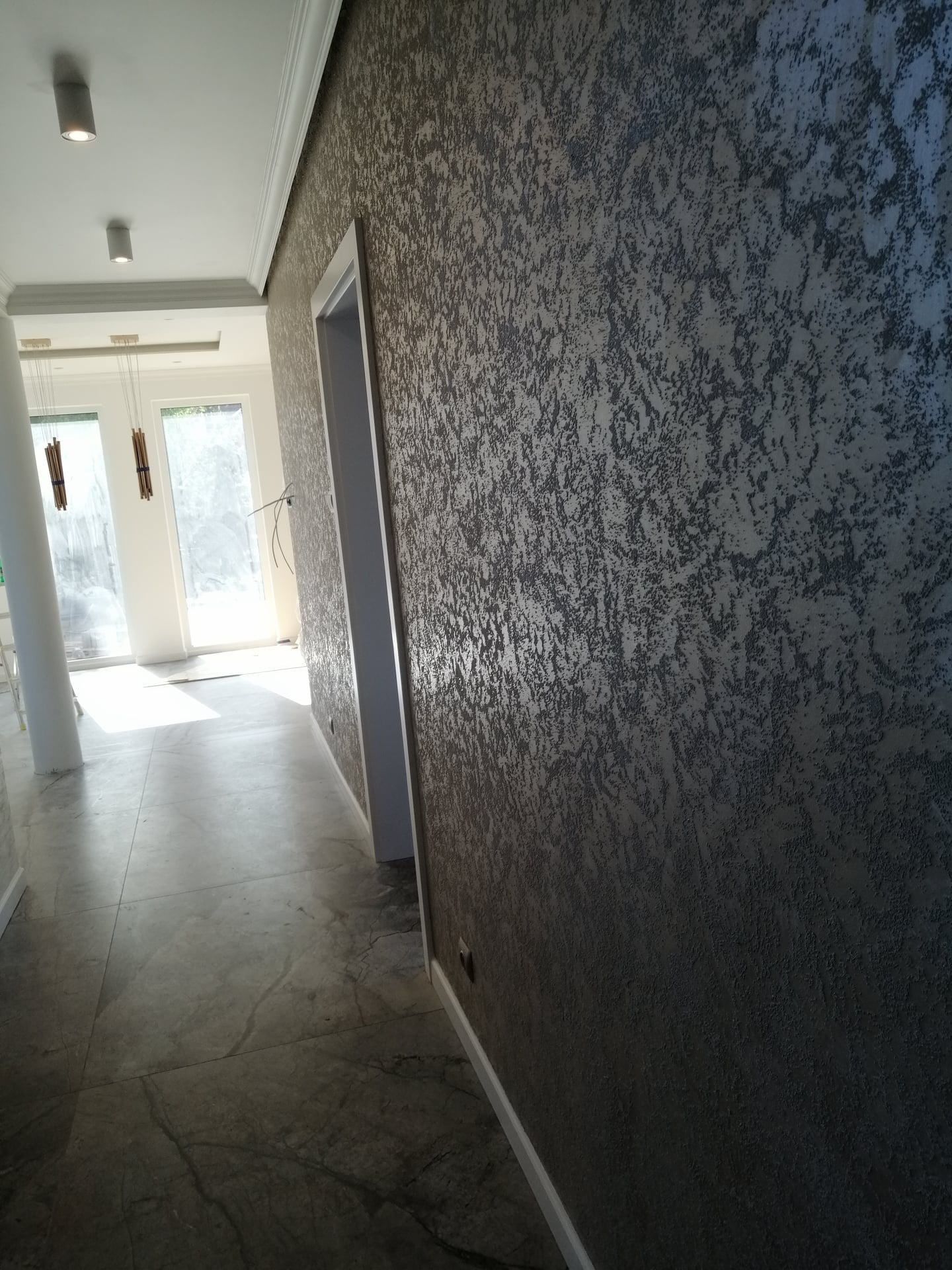 Korytarz z dekoracyjną, ciemną, strukturalną ścianą, marmurową podłogą i białymi futrynami drzwi oraz widokiem na jasne pomieszczenie z oknami.