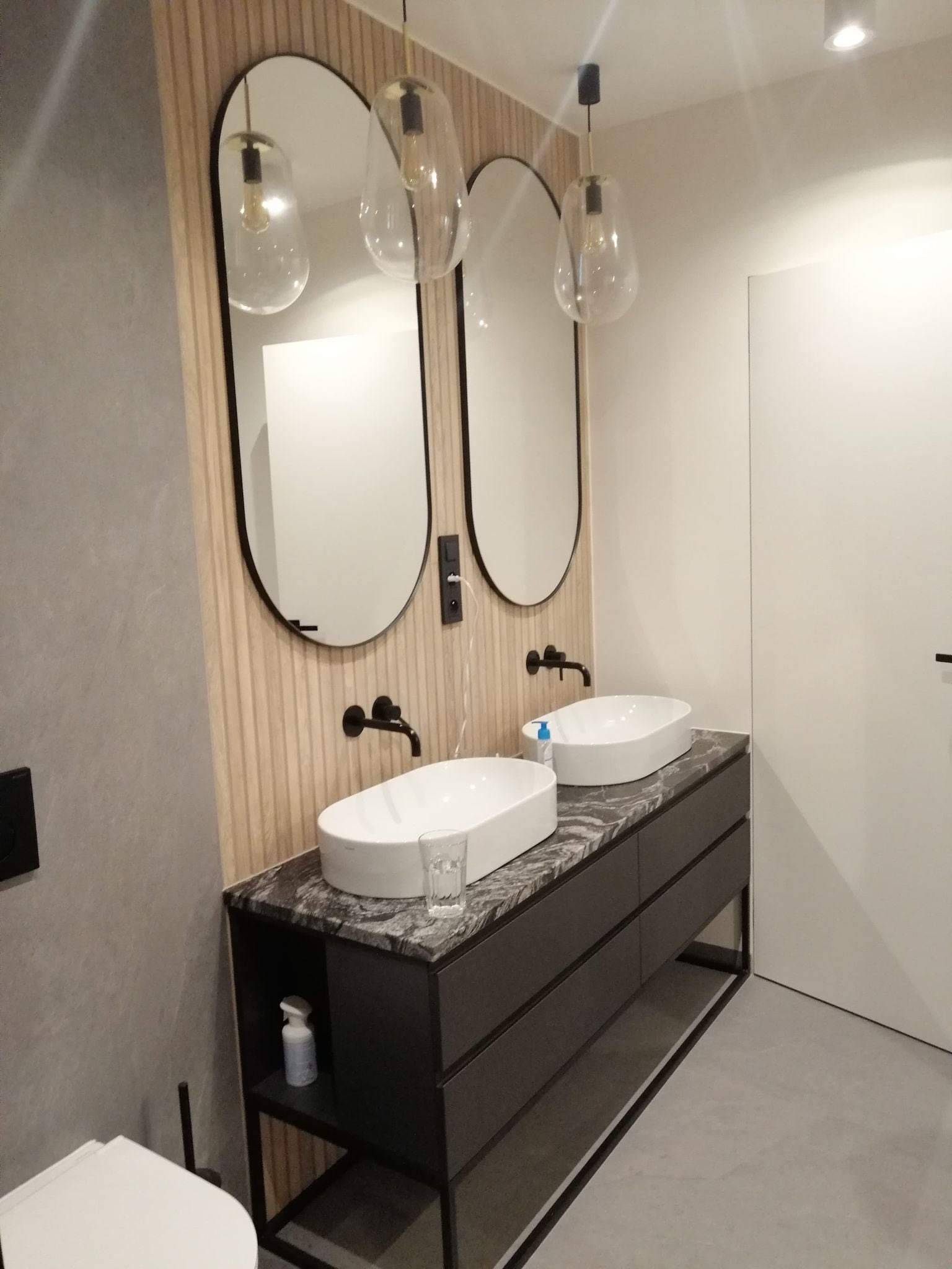 Nowoczesna łazienka z dwoma owalnymi umywalkami nablatowymi, czarną armaturą, lustrami w czarnych ramach i wiszącymi lampami z żarówkami Edisona, ściana wykończona panelami drewnopodobnymi.