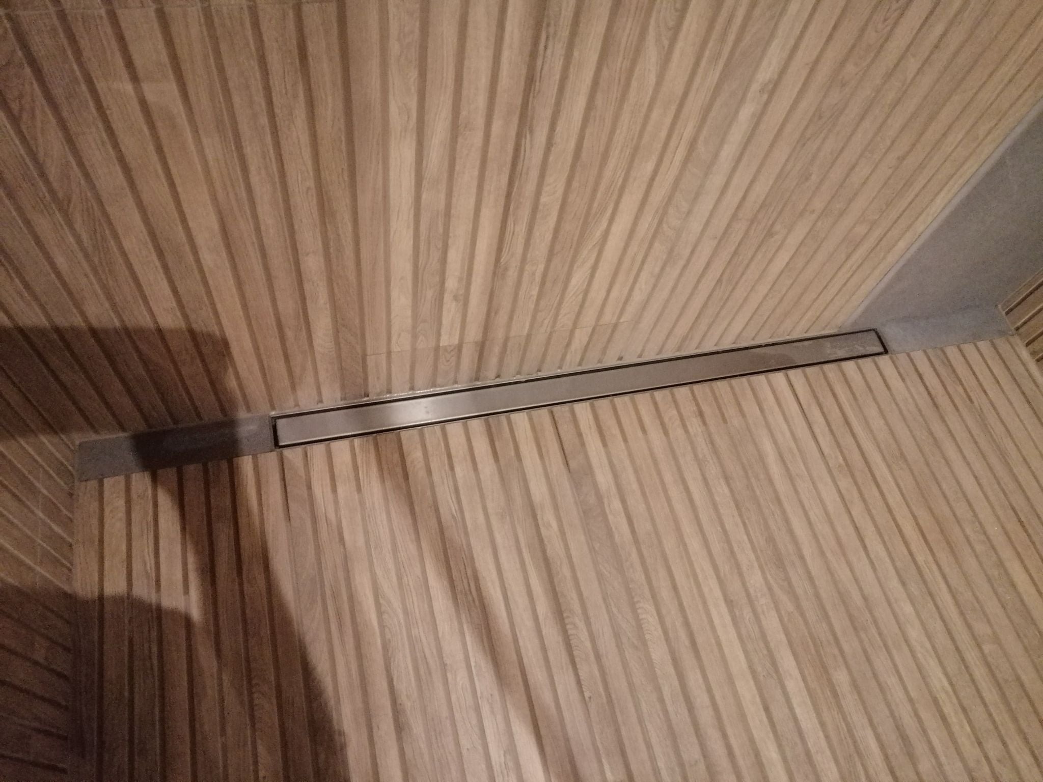 Ściana i podłoga wykończone panelami z drewna o pionowym wzorze, z widocznym liniowym odpływem prysznicowym w metalowej ramie.