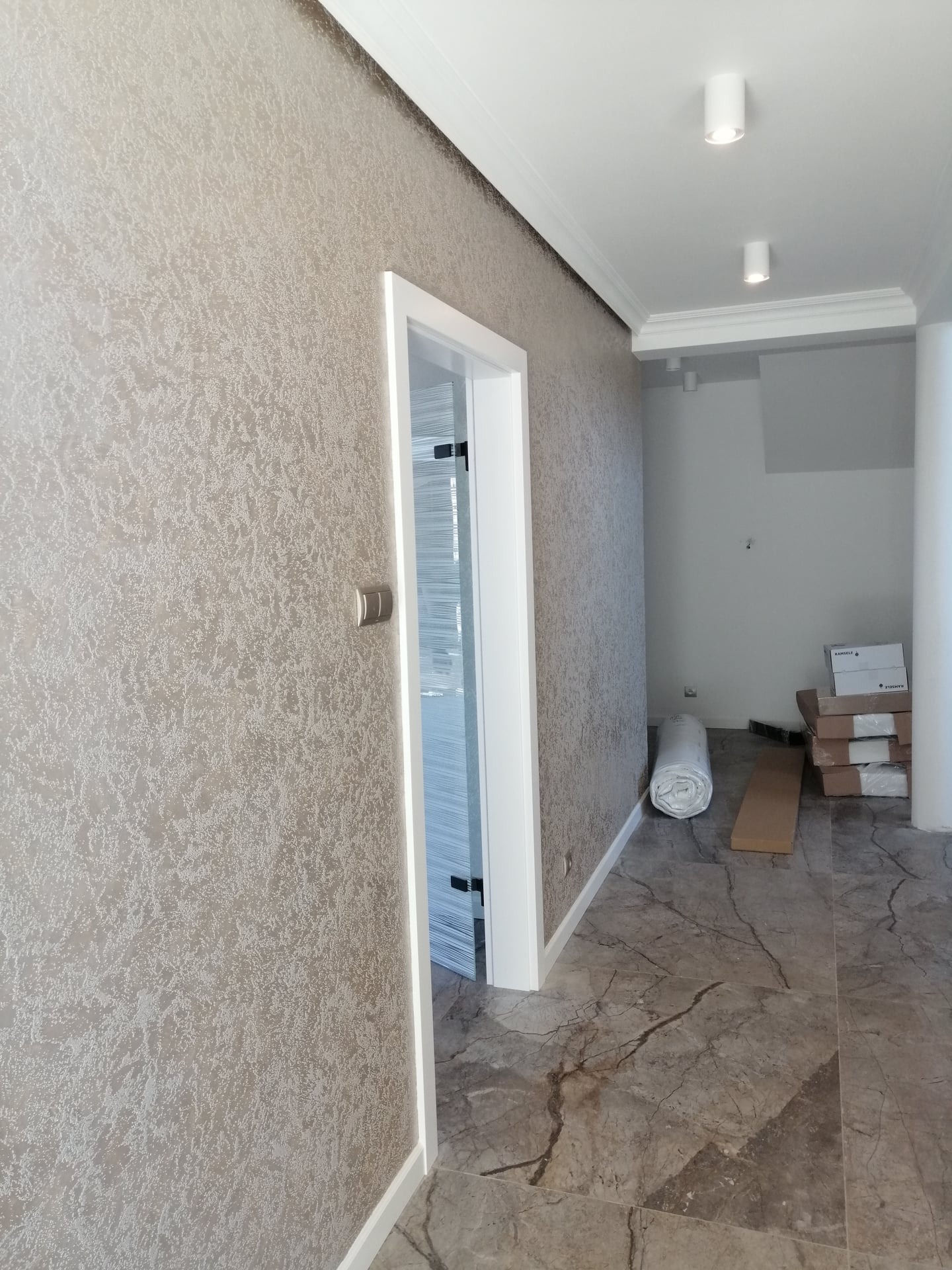 Ściana z beżową, teksturowaną tapetą przy białych drzwiach, marmurowa podłoga, biały sufit z oświetleniem punktowym i dekoracyjną listwą przysufitową; w głębi pokoju zwinięty dywan i kartony.