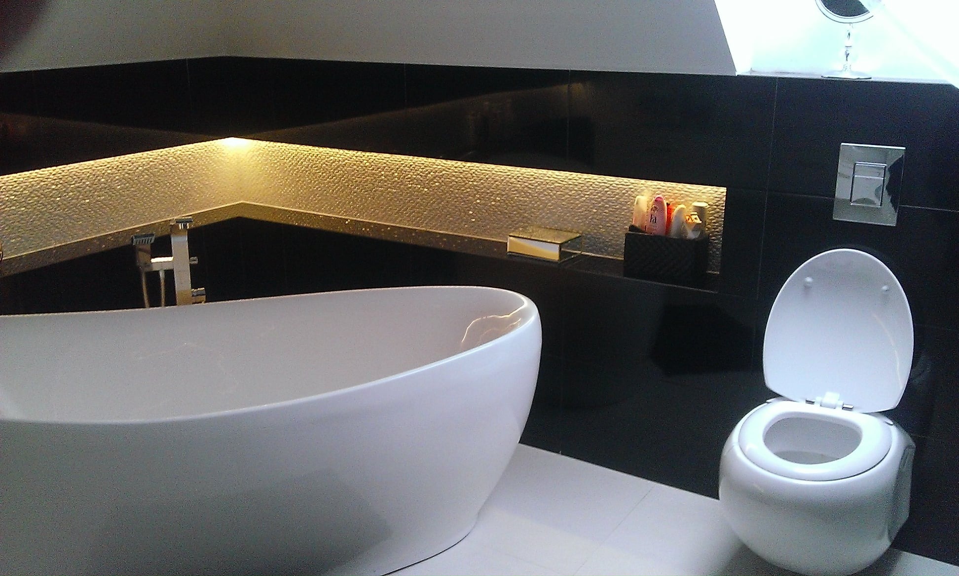 Nowoczesna łazienka z wolnostojącą wanną, podświetlaną wnęką z mozaiką i wiszącą toaletą, wykończona czarnymi płytkami i białymi fugami.