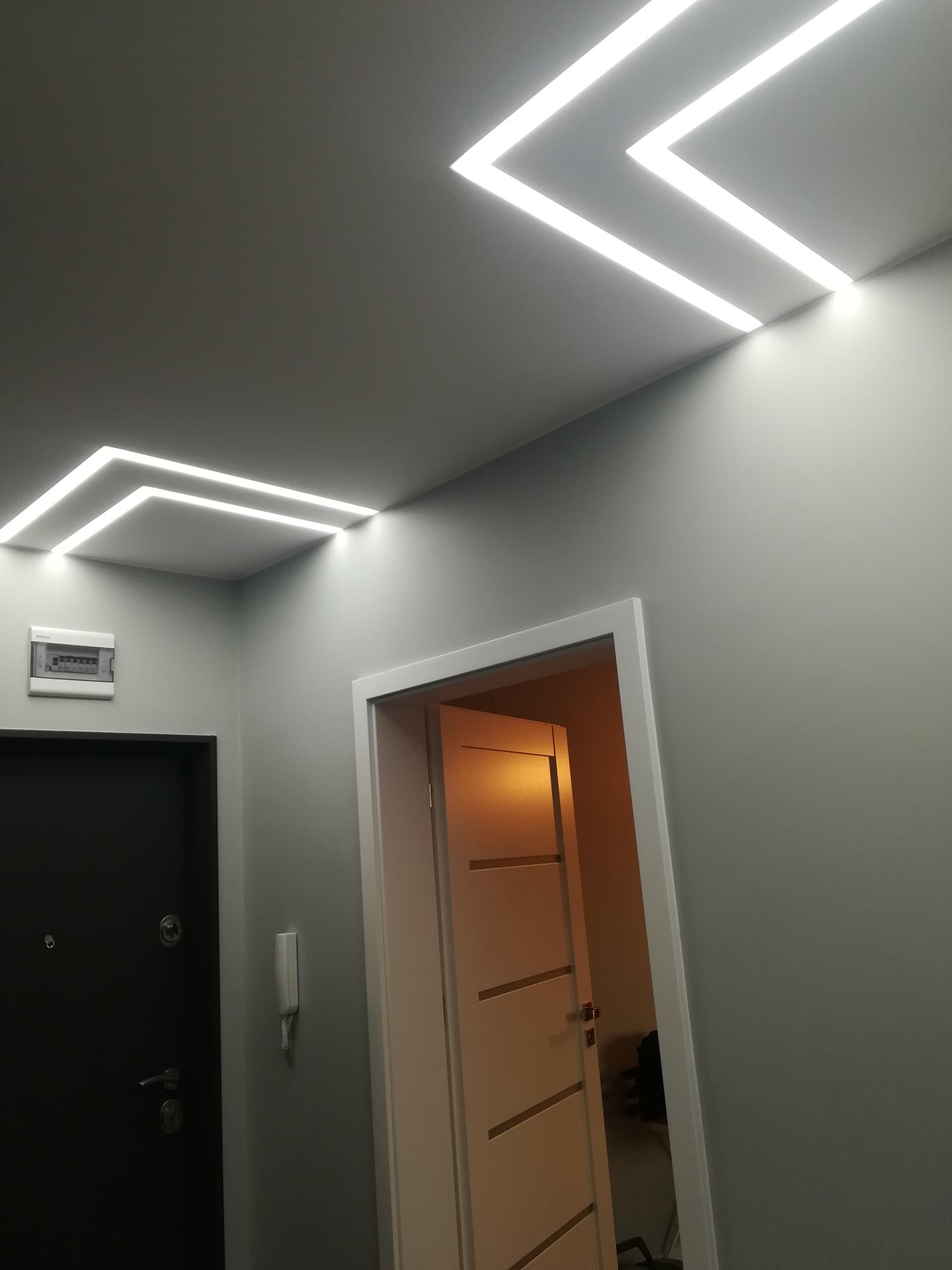 Korytarz z nowoczesnym oświetleniem sufitowym LED w kształcie geometrycznym, szare ściany, czarne drzwi wejściowe i otwarte białe drzwi wewnętrzne.