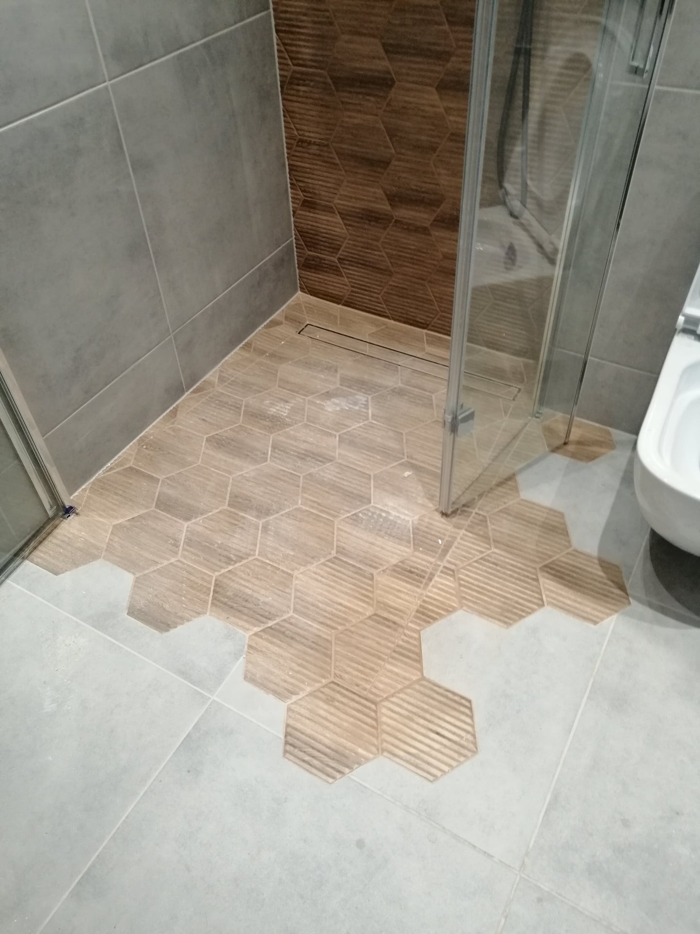 Ułożone heksagonalne płytki drewnopodobne w kabinie prysznicowej, przechodzące w szare płytki podłogowe, z widocznym odpływem liniowym i częścią toalety.