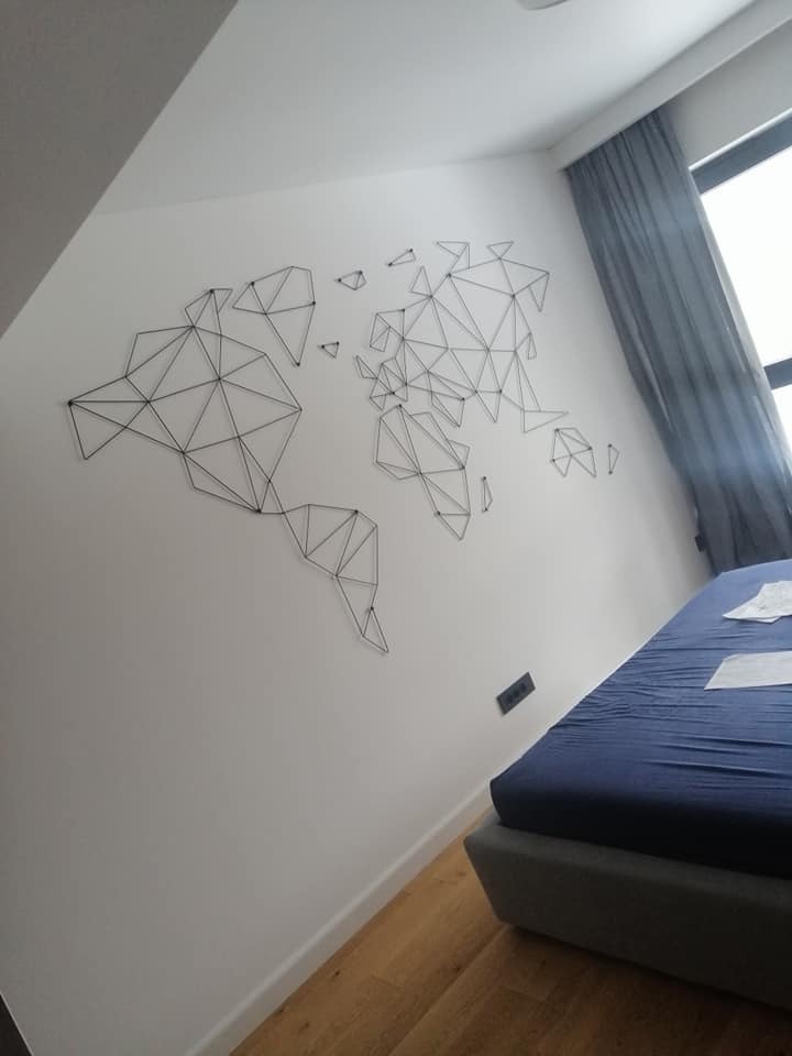 Geometryczna mapa świata z czarnego drutu na białej ścianie w sypialni z drewnianą podłogą i zasłoniętym oknem.