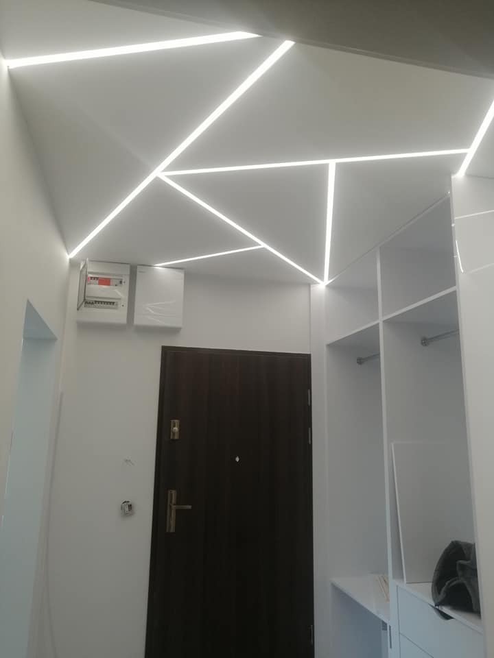 Nowoczesny przedpokój z geometrycznym oświetleniem LED na suficie, brązowe drzwi wejściowe, biała zabudowa garderoby i skrzynka elektryczna na ścianie.