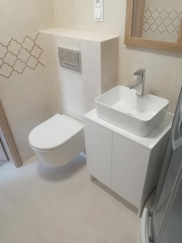 Nowoczesna łazienka w jasnych barwach z podwieszaną toaletą, umywalką nablatową na szafce i dekoracyjną mozaiką na ścianie, widok z boku.