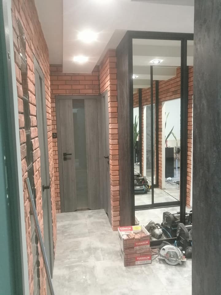 Korytarz w Łodzi z wykończeniem ścian imitującym cegłę, szara podłoga, szare drzwi z przeszkleniami i czarna konstrukcja szklanej ściany. Na podłodze widoczne narzędzia i opakowania materiałów...