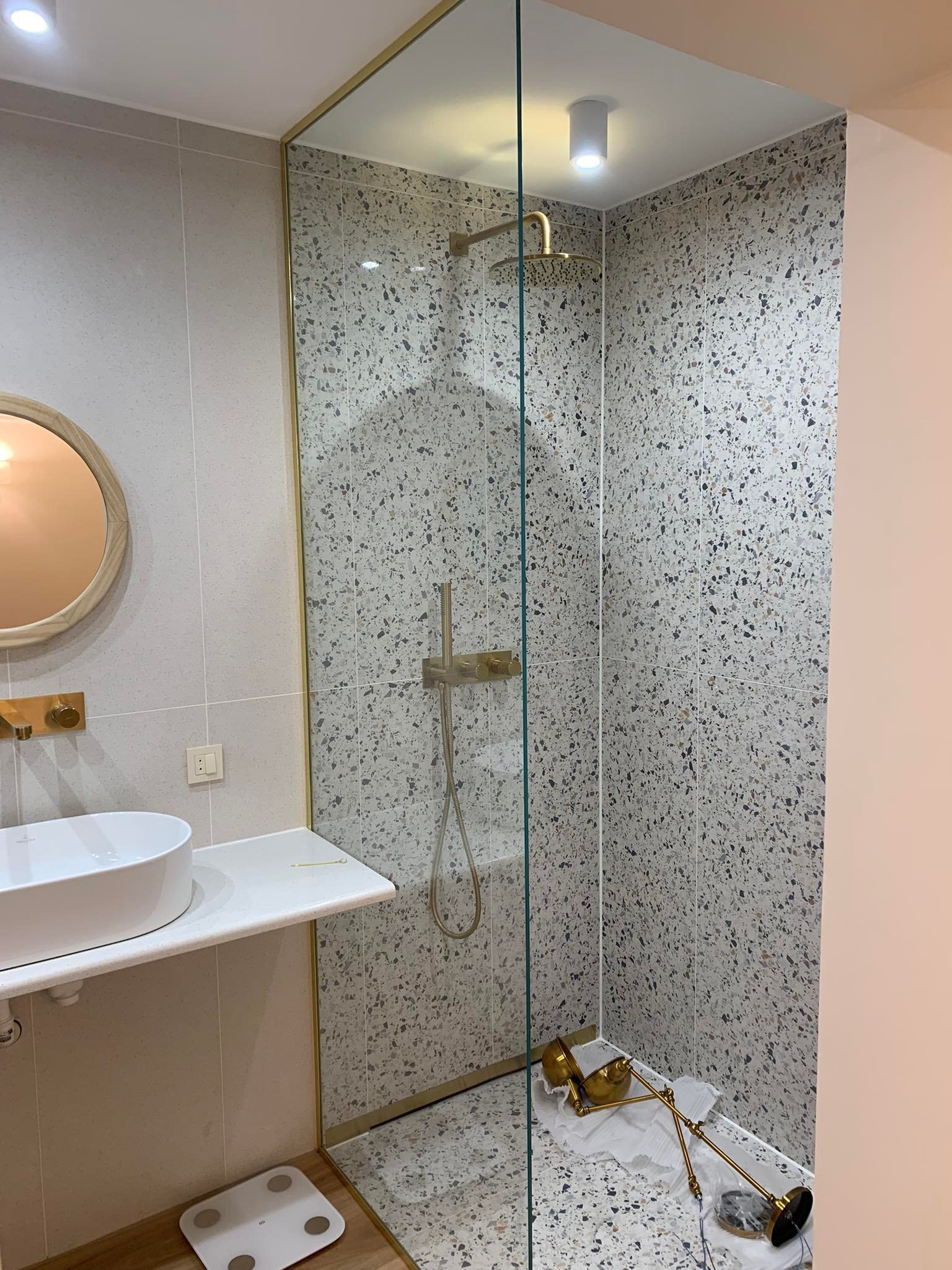 Nowoczesna łazienka z kabiną prysznicową typu walk-in, wykończona płytkami imitującymi lastryko, z elementami armatury w kolorze złotym i białym umywalką nablatową, oraz wagą łazienkową na podłodze...