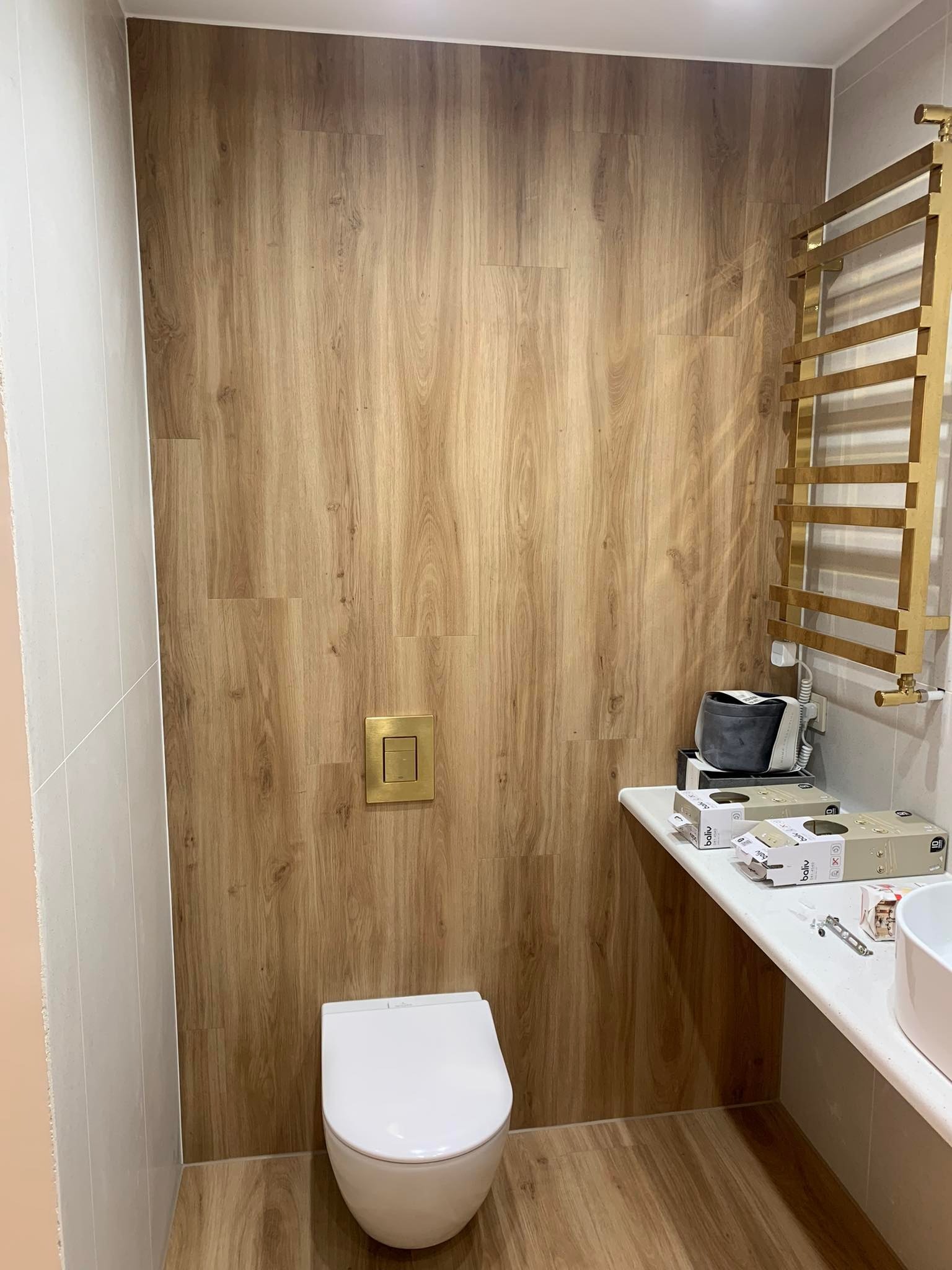 Wykończona łazienka z panelami imitującymi drewno na ścianach i podłodze, złoty grzejnik drabinkowy, biała toaleta podwieszana z złotym przyciskiem spłukiwania, umywalka nablatowa i akcesoria...