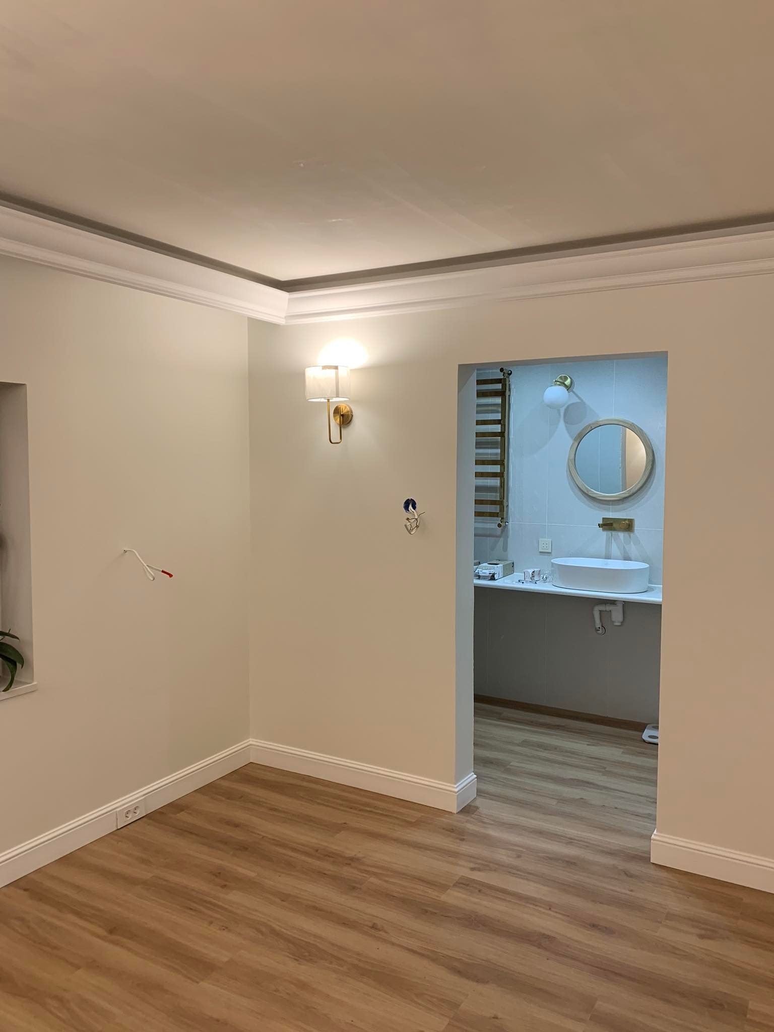 Jasne wnętrze po remoncie: drewnopodobna podłoga, świeżo pomalowane ściany w odcieniach beżu, dekoracyjne listwy przypodłogowe i sufitowe, widoczne gniazdka elektryczne i fragment łazienki...