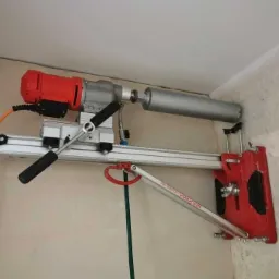 Wiertnica diamentowa z czerwonym silnikiem i srebrnym wiertłem przymocowana do ściany, widoczne przewody i elementy montażowe.