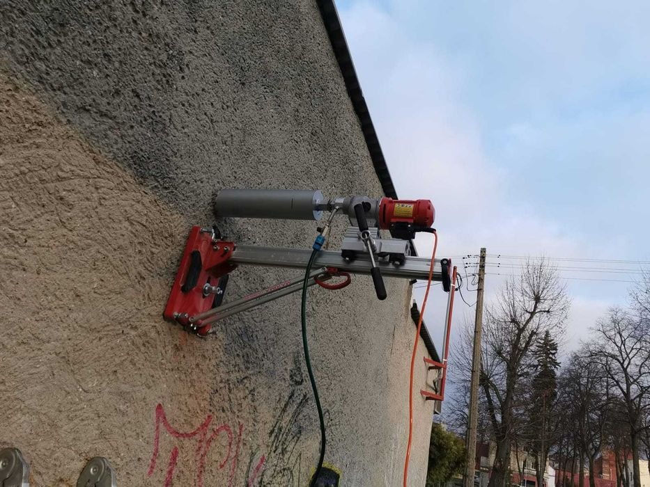 Wiertnica diamentowa przymocowana do ściany budynku podczas wykonywania otworu, widoczne przewody i elementy mocujące, tło stanowi fragment nieba i drzewa.