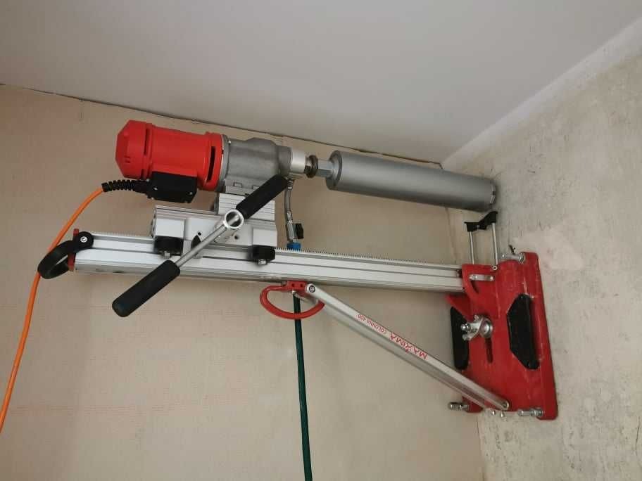 Wiertnica diamentowa z czerwonym silnikiem i srebrnym wiertłem przymocowana do ściany, widoczne przewody i elementy montażowe.