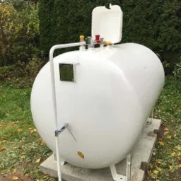 Biały zbiornik na gaz płynny umieszczony na betonowej podstawie w ogrodzie, widoczne zawory i rura doprowadzająca gaz do ziemi.