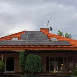 Dom jednorodzinny z czerwoną dachówką i panelami słonecznymi zamontowanymi na dachu. Widoczne okna dachowe, antena satelitarna i komin. Ogród z różnorodną roślinnością.