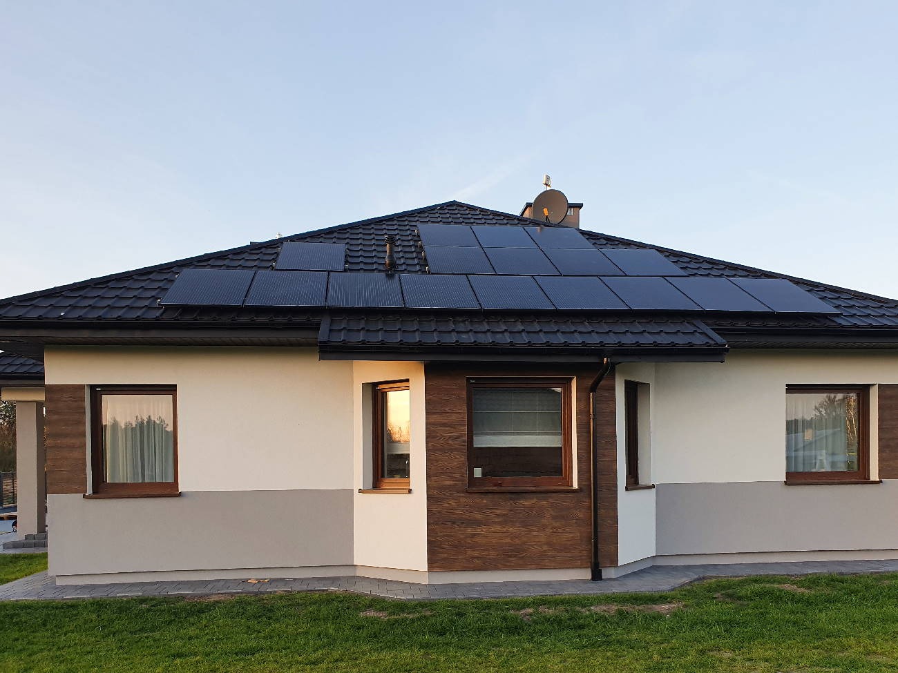 Dom jednorodzinny z panelami słonecznymi na dachu z blachodachówki, widoczne okna z drewnianymi ramami i fragment trawnika przed budynkiem.