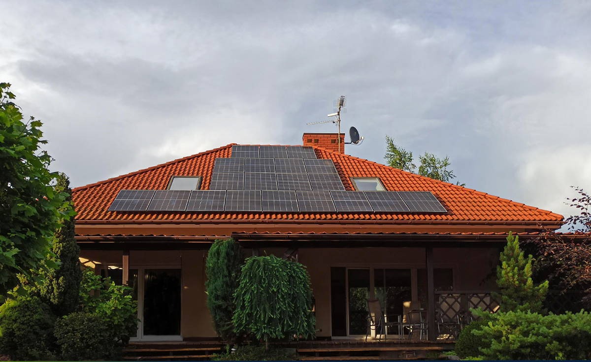 Dom jednorodzinny z czerwoną dachówką i panelami słonecznymi zamontowanymi na dachu. Widoczne okna dachowe, antena satelitarna i komin. Ogród z różnorodną roślinnością.