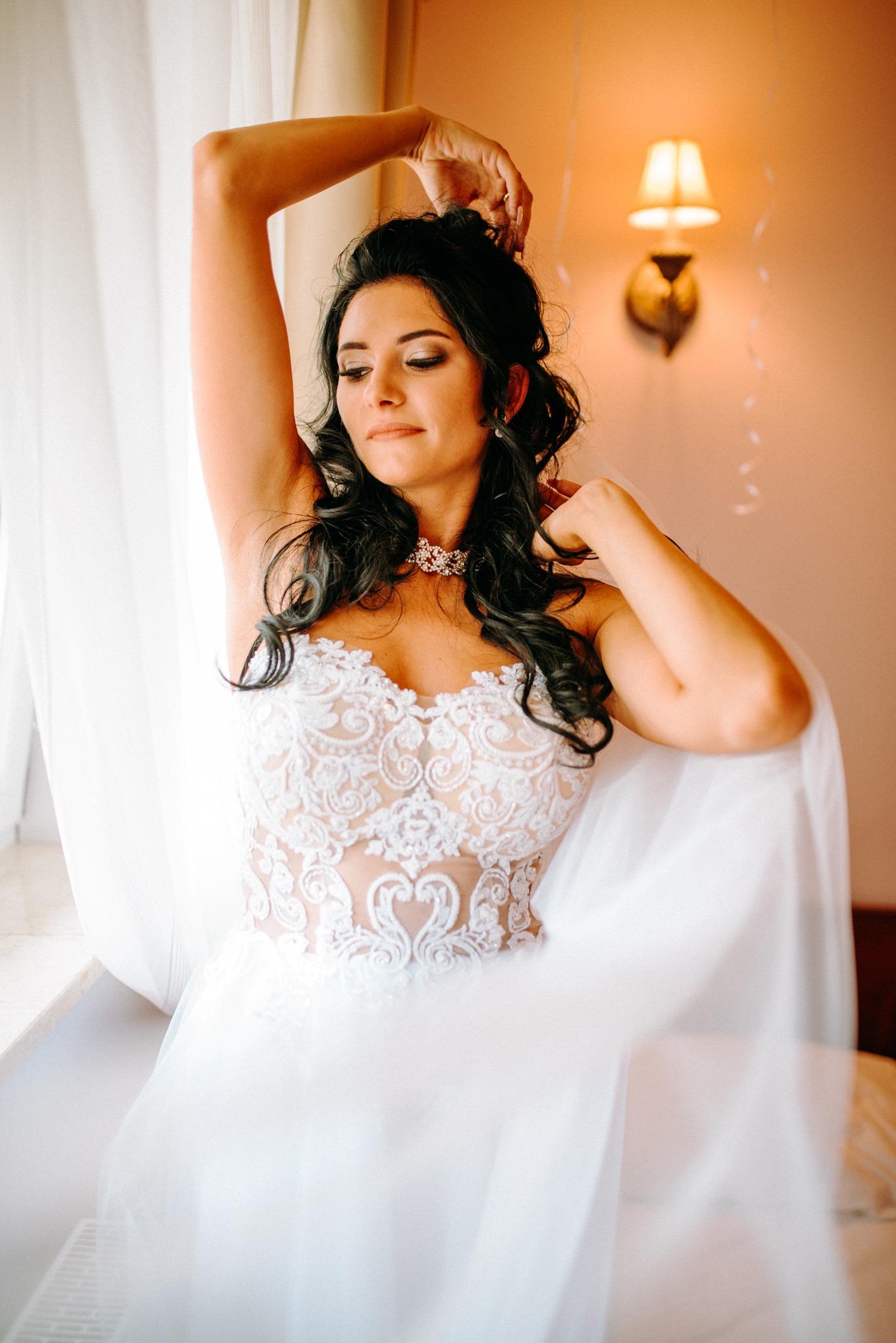 Panna młoda w koronkowej sukni, pozująca przy oknie z delikatnym światłem, z upiętymi włosami i subtelnym makijażem, ozdobiona błyszczącym naszyjnikiem.