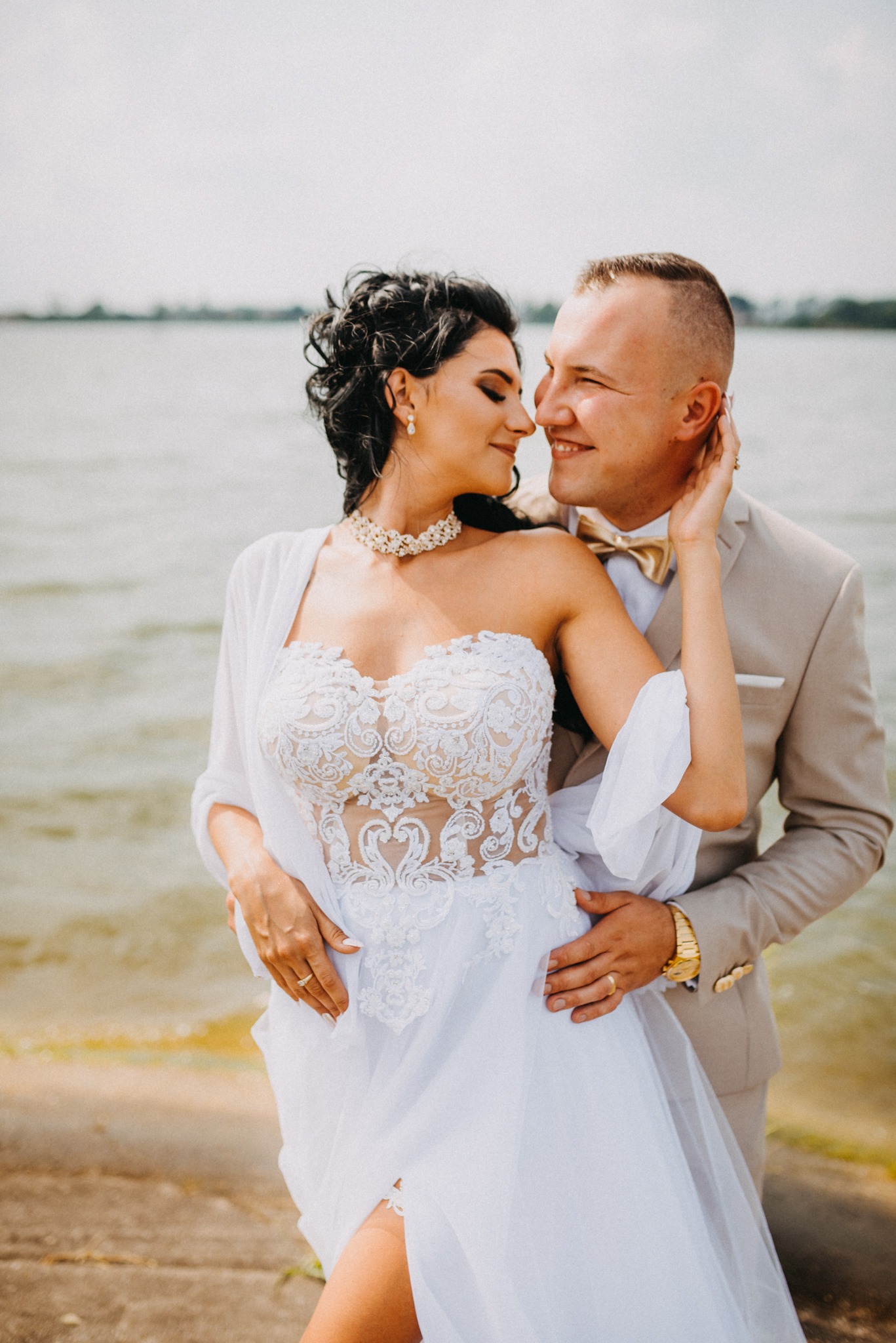 Para młoda w objęciach na tle jeziora: panna młoda w koronkowej sukni bez ramiączek i chokerze, pan młody w beżowym garniturze i złotej muszce.