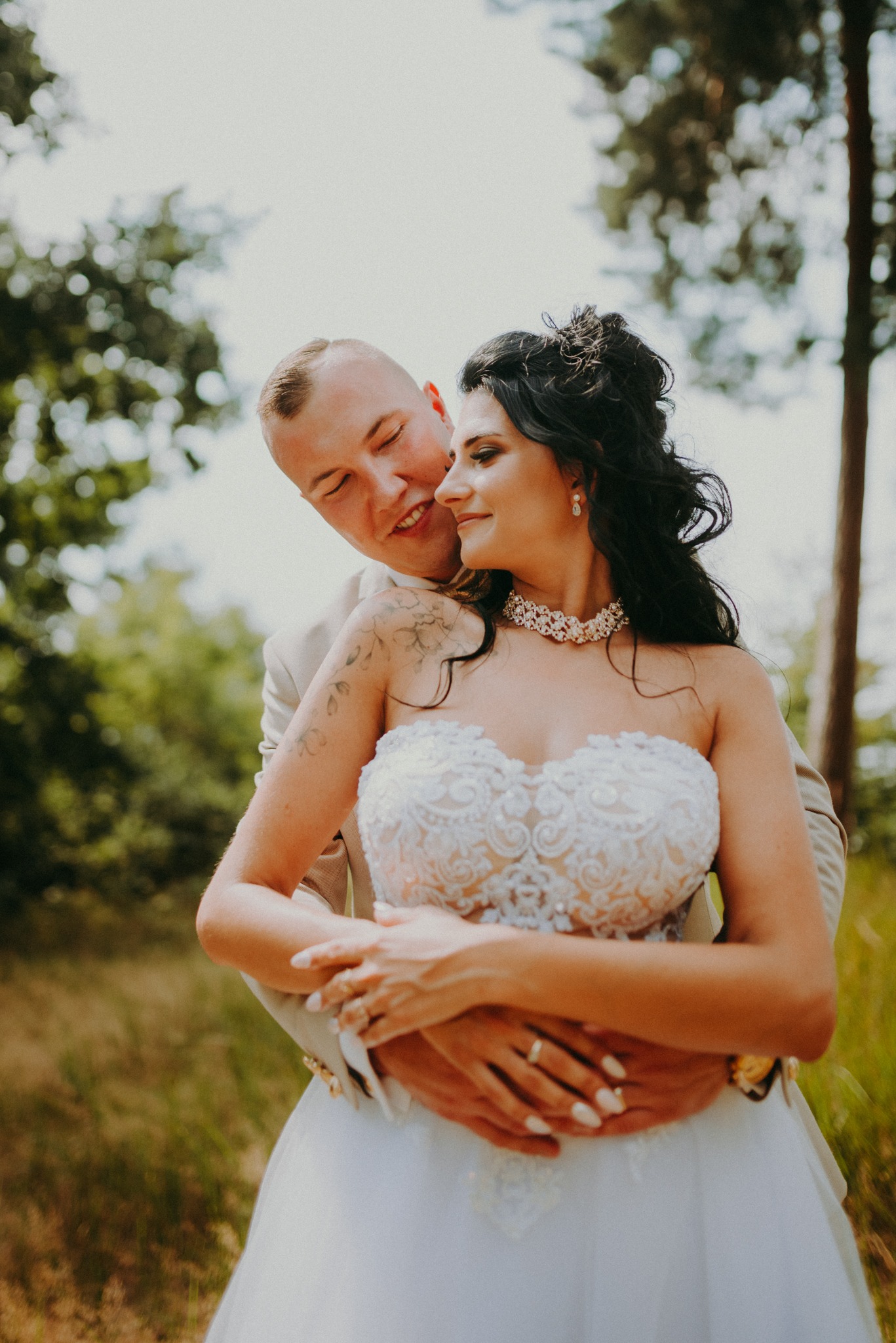 Para młoda w objęciach na tle drzew: panna młoda w koronkowej sukni bez ramiączek i z naszyjnikiem, pan młody w beżowym garniturze z tatuażem na ramieniu.