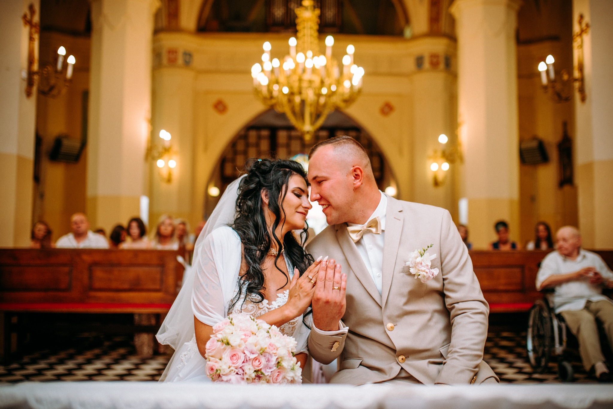 Para młoda w kościele, zbliżona twarzami, prezentująca obrączki na tle żyrandola i zgromadzonych gości.
