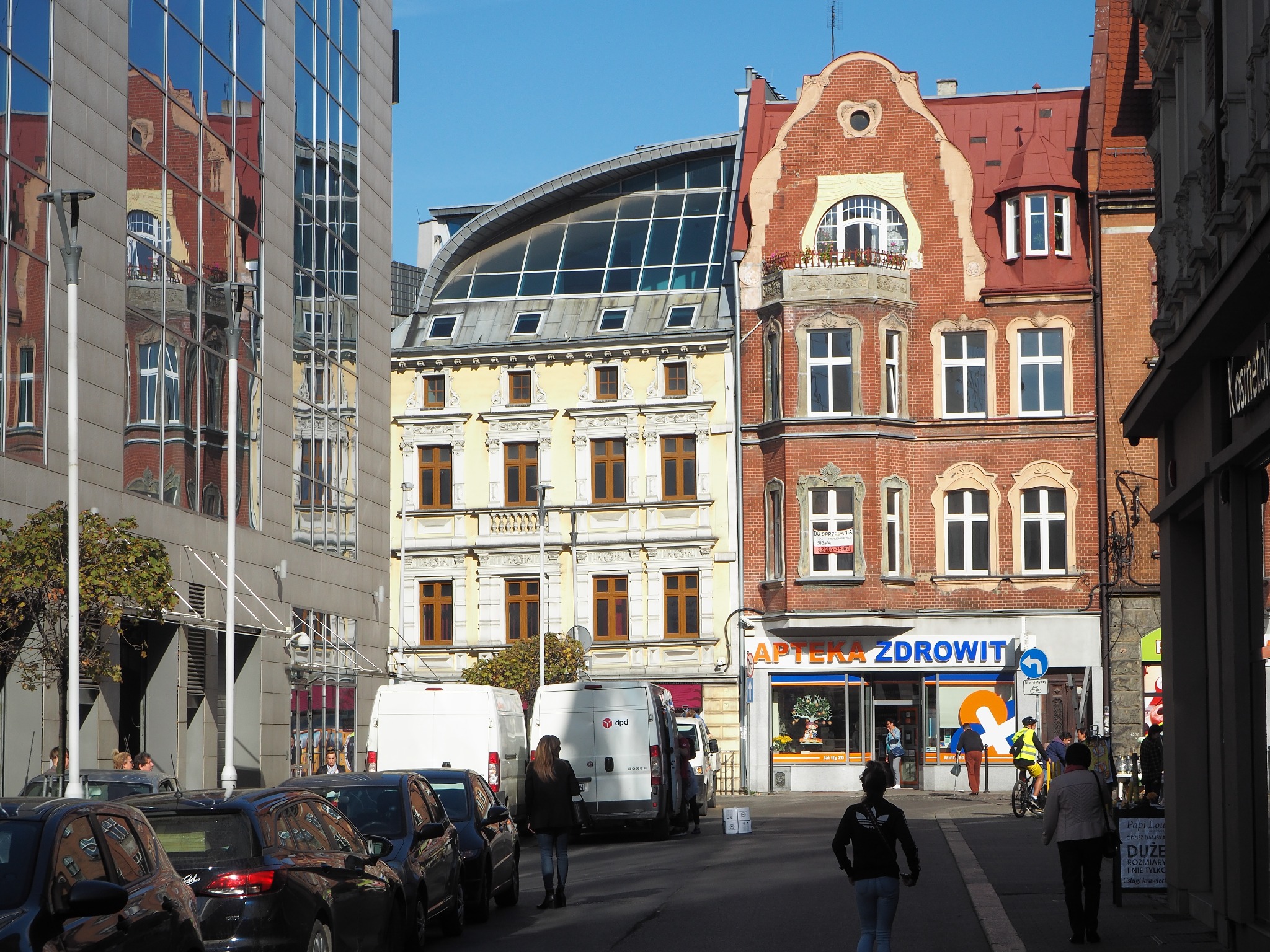 Ulica w Bytomiu z różnorodną architekturą: nowoczesny biurowiec odbija kamienice, a na chodniku parkują samochody dostawcze i spacerują przechodnie.