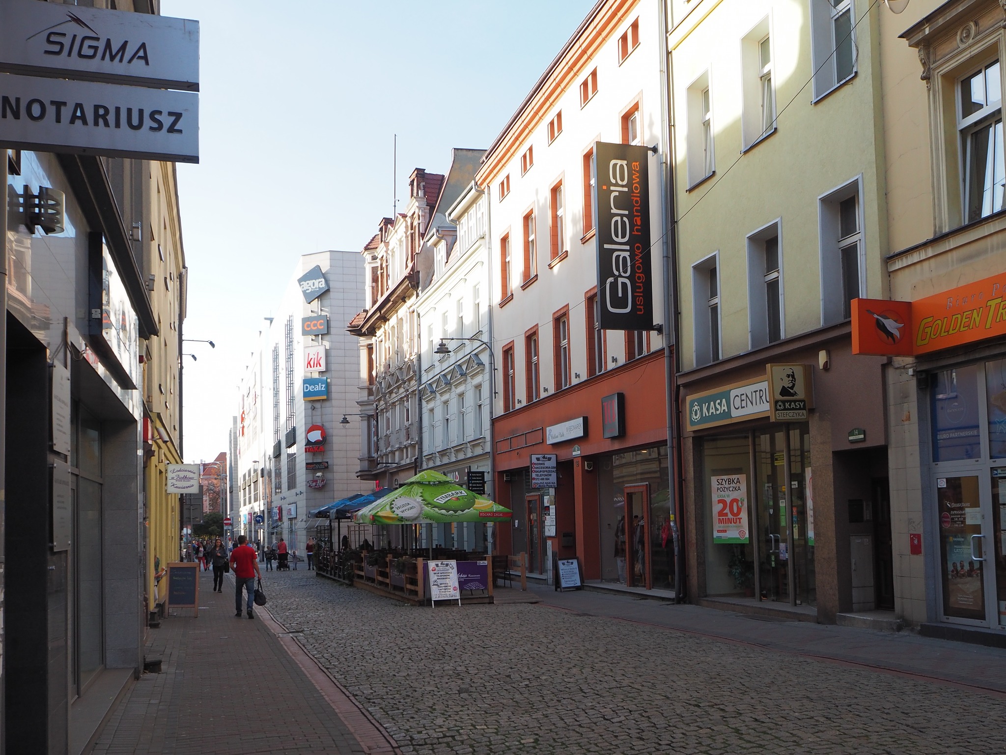 Ulica w centrum miasta z budynkami biurowymi i usługowymi, w tle widoczne szyldy firm, na pierwszym planie brukowana ulica i chodnik z przechodniami.