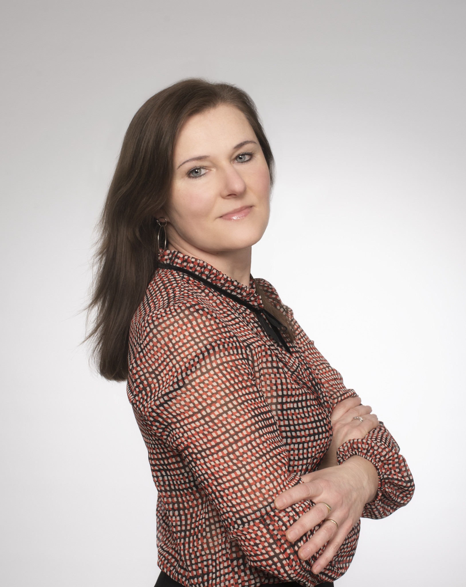 Portret kobiety z brązowymi włosami, ubranej w bluzkę w geometryczny wzór, z rękami skrzyżowanymi na piersiach, na jasnym tle.