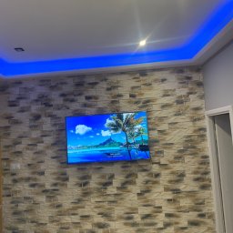 PatKoz - Ściana wykończona dekoracyjną okładziną imitującą kamień, z zawieszonym telewizorem wyświetlającym krajobraz tropikalny; sufit z podświetleniem LED w kolorze niebieskim.
