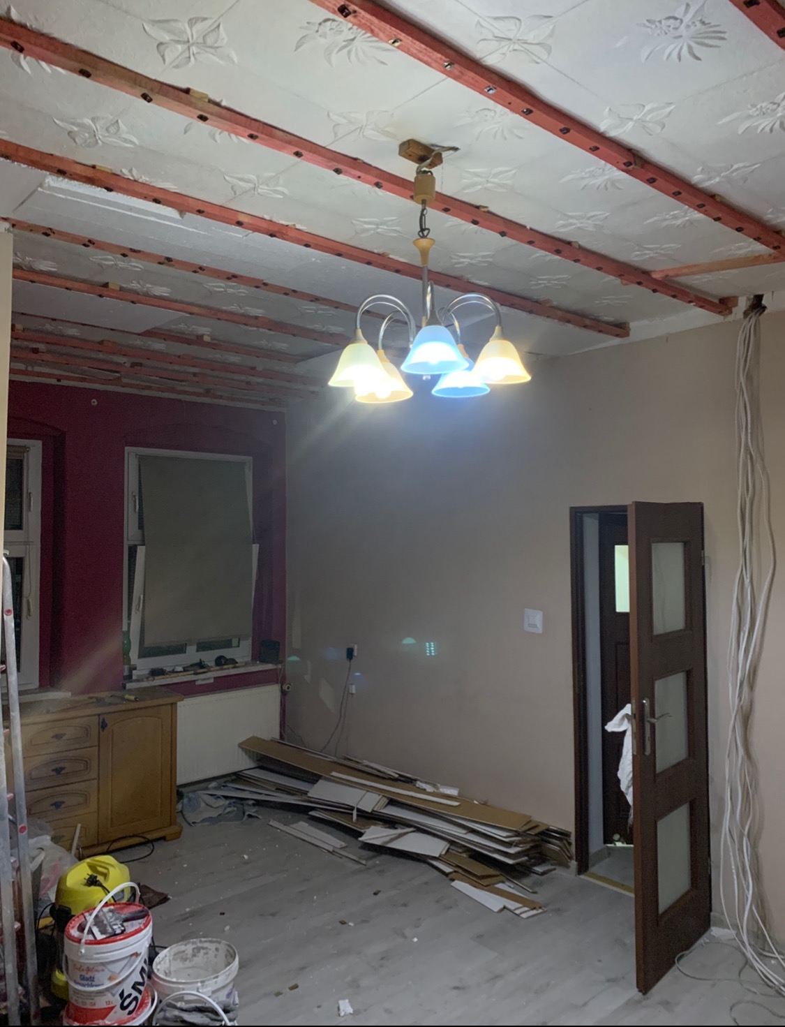 Wnętrze w trakcie remontu, widoczne drewniane legary przytwierdzone do sufitu z dekoracyjnym wzorem, wiszący żyrandol z kolorowymi kloszami, podłoga częściowo zakryta panelami, ściany w dwóch...