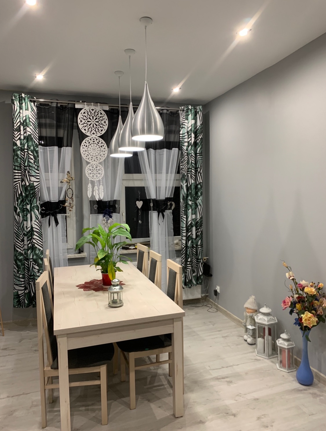 Jasne wnętrze jadalni z drewnianym stołem i krzesłami, ozdobione roślinami doniczkowymi i lampionami, podwieszane lampy w kształcie kropli, wzorzyste zasłony w oknach.