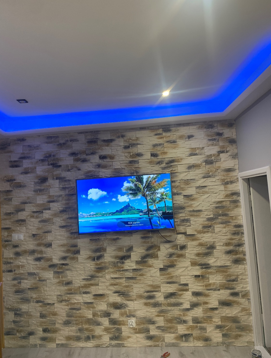 Ściana wykończona dekoracyjną okładziną imitującą kamień, z zawieszonym telewizorem wyświetlającym krajobraz tropikalny; sufit z podświetleniem LED w kolorze niebieskim.
