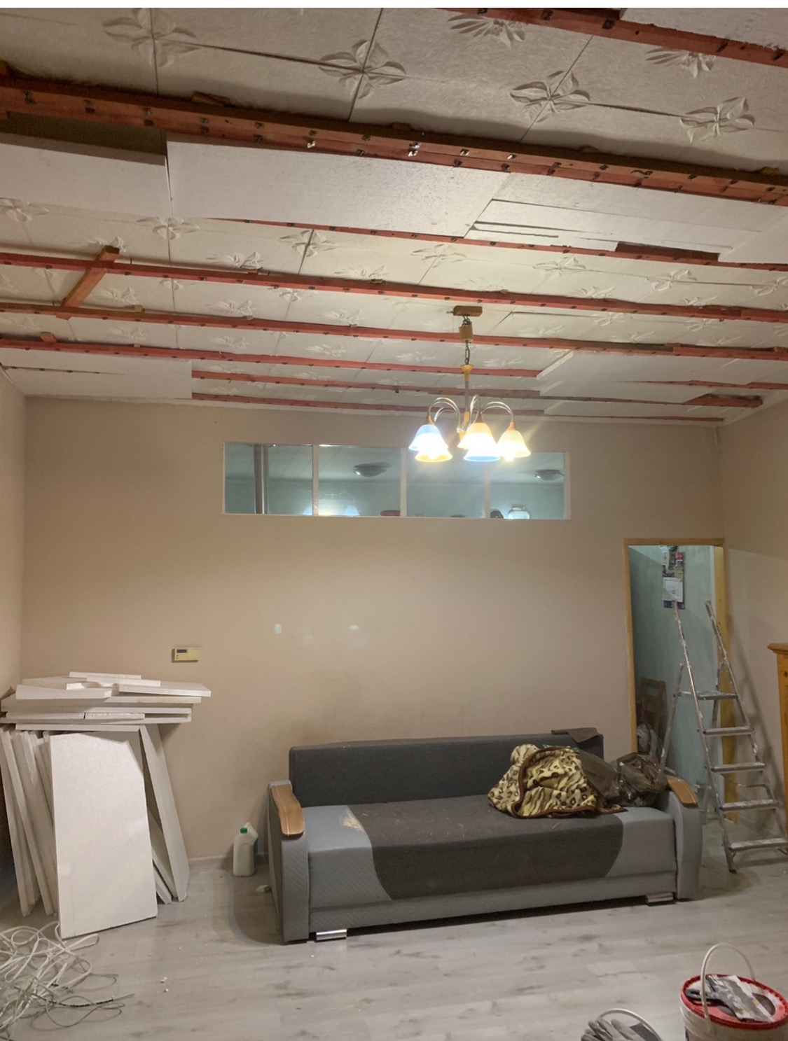 Wnętrze w trakcie prac wykończeniowych: częściowo zamontowane płyty sufitowe z dekoracyjnym wzorem, drewniana konstrukcja podtrzymująca, sofa w pokoju, oraz materiały budowlane w rogu pomieszczenia.