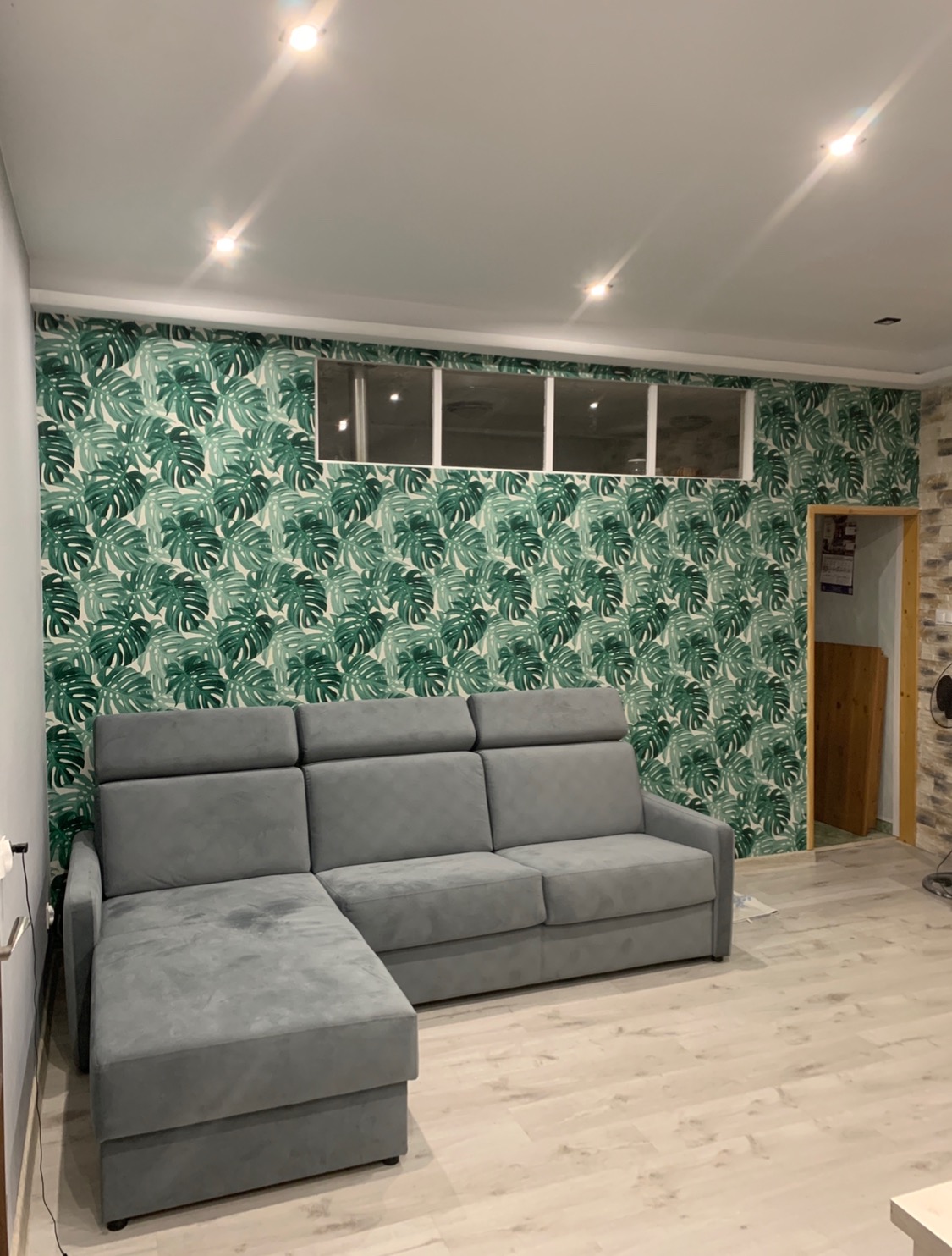 Szary narożnik z regulowanymi zagłówkami na tle ściany pokrytej tapetą w zielone liście monstery, trzy okna pod sufitem, jasna podłoga.