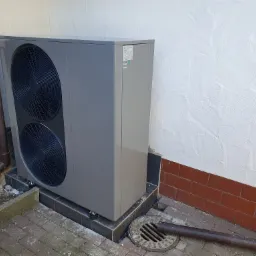 Szara pompa ciepła powietrze-woda zainstalowana przy ścianie budynku z widocznym odpływem wody deszczowej