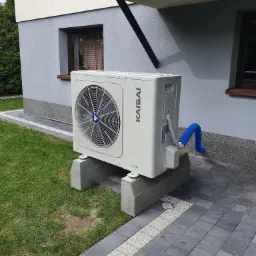 Klimatyzator Kaisai zamontowany na betonowych blokach przy budynku w Woli Mysłowskiej, widoczny niebieski wąż odprowadzający skropliny.