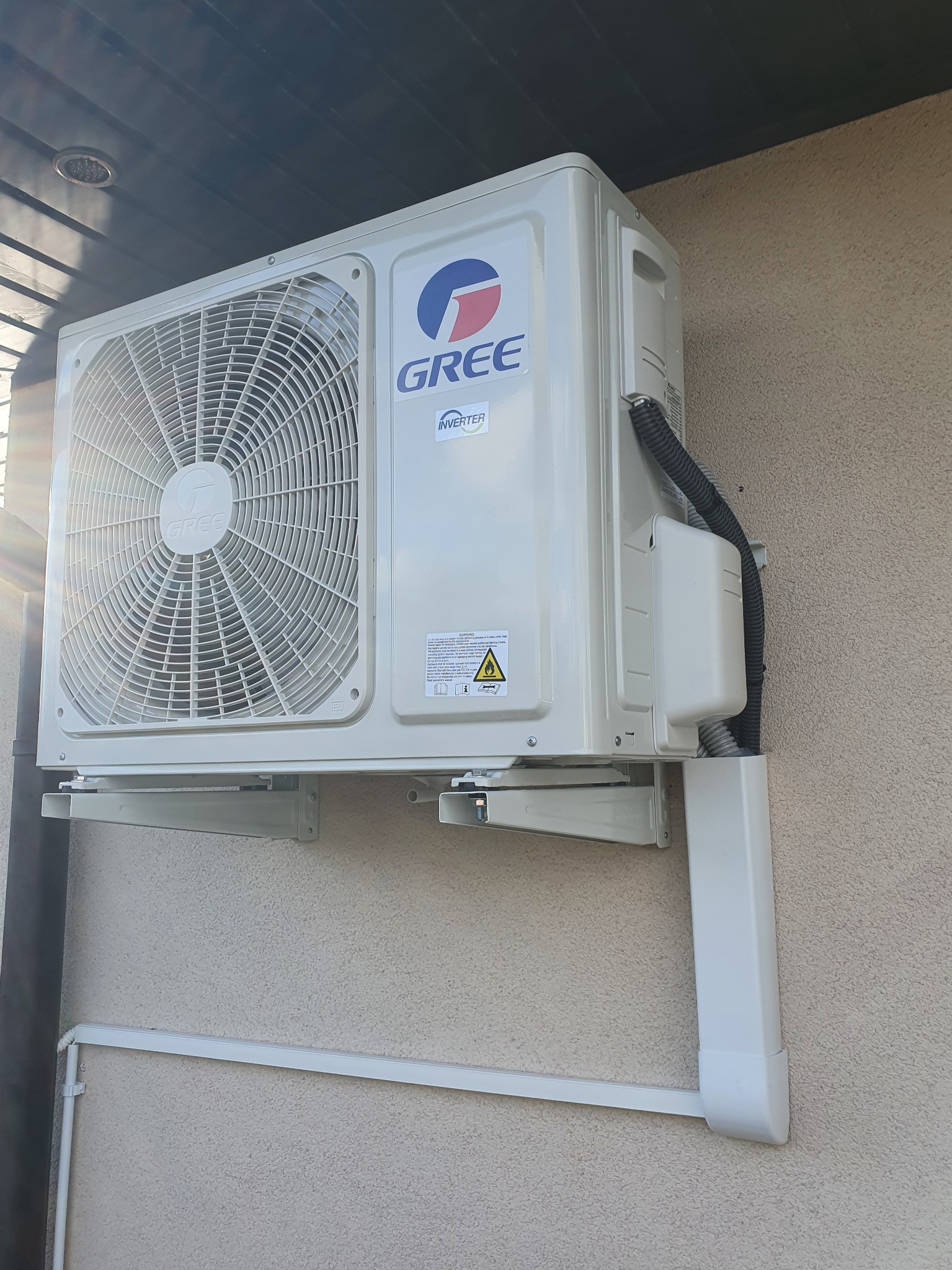 Zewnętrzna jednostka klimatyzacji marki Gree zamontowana na elewacji budynku, widoczne przewody i rurki odprowadzające skropliny.