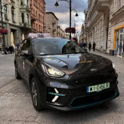 Czarny samochód taxi Kia Niro z podświetlanymi elementami w kolorze turkusowym, zaparkowany na brukowanej ulicy w centrum miasta, widok z przodu, tablica rejestracyjna WI 046LR.