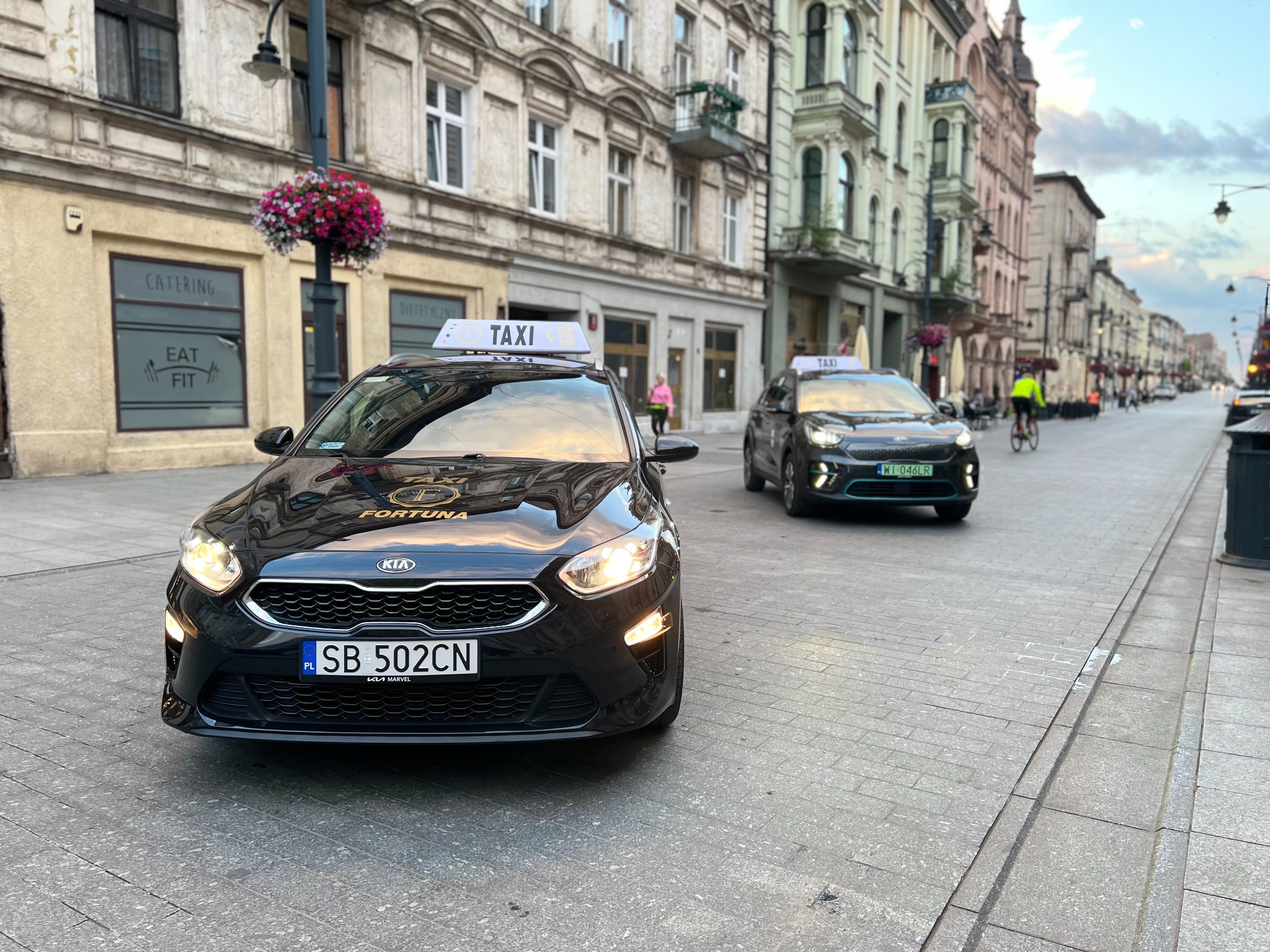 Dwa czarne samochody taxi marki Kia zaparkowane na brukowanej ulicy w centrum miasta, z widocznymi tablicami rejestracyjnymi i napisem 'FORTUNA' na masce pierwszego pojazdu.