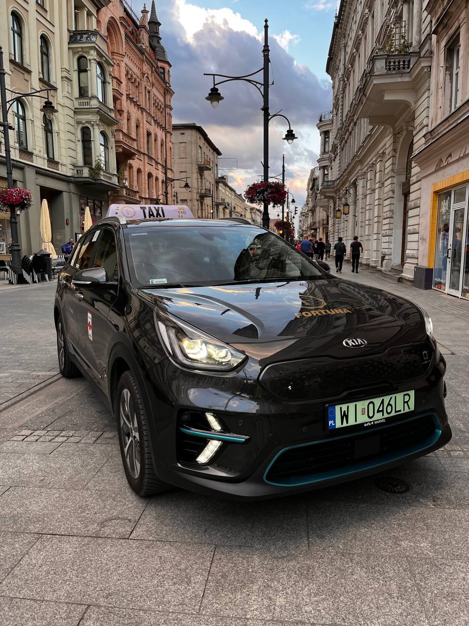 Czarny samochód taxi Kia Niro z podświetlanymi elementami w kolorze turkusowym, zaparkowany na brukowanej ulicy w centrum miasta, widok z przodu, tablica rejestracyjna WI 046LR.