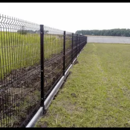 Długi, prosty odcinek czarnego, panelowego ogrodzenia z podmurówką, oddzielający pole z trawą od zaoranej ziemi, z budynkiem w tle.