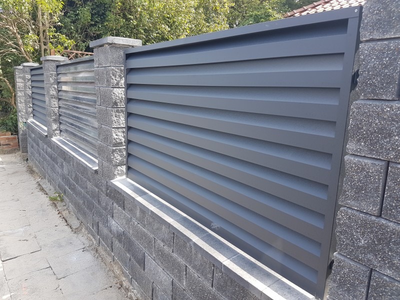 Nowoczesne ogrodzenie z poziomych, szarych paneli zamontowanych na murku z szarej kostki betonowej, z elementami przeziernymi.