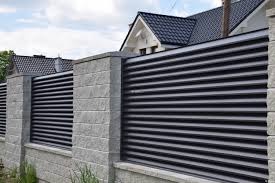 Nowoczesne ogrodzenie panelowe z poziomych, czarnych profili zamontowane na słupkach z szarej kostki betonowej, w tle dom z ciemną dachówką.