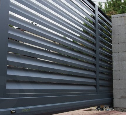 Nowoczesna brama przesuwna z poziomymi lamelami w kolorze grafitowym, widoczny fragment muru z bloczków betonowych.