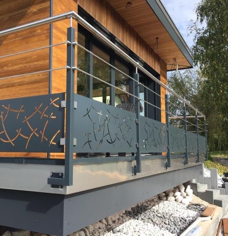 Nowoczesna balustrada balkonowa z metalowymi panelami dekoracyjnymi i stalowymi poręczami, zamontowana przy drewnianym domu z dużymi oknami.