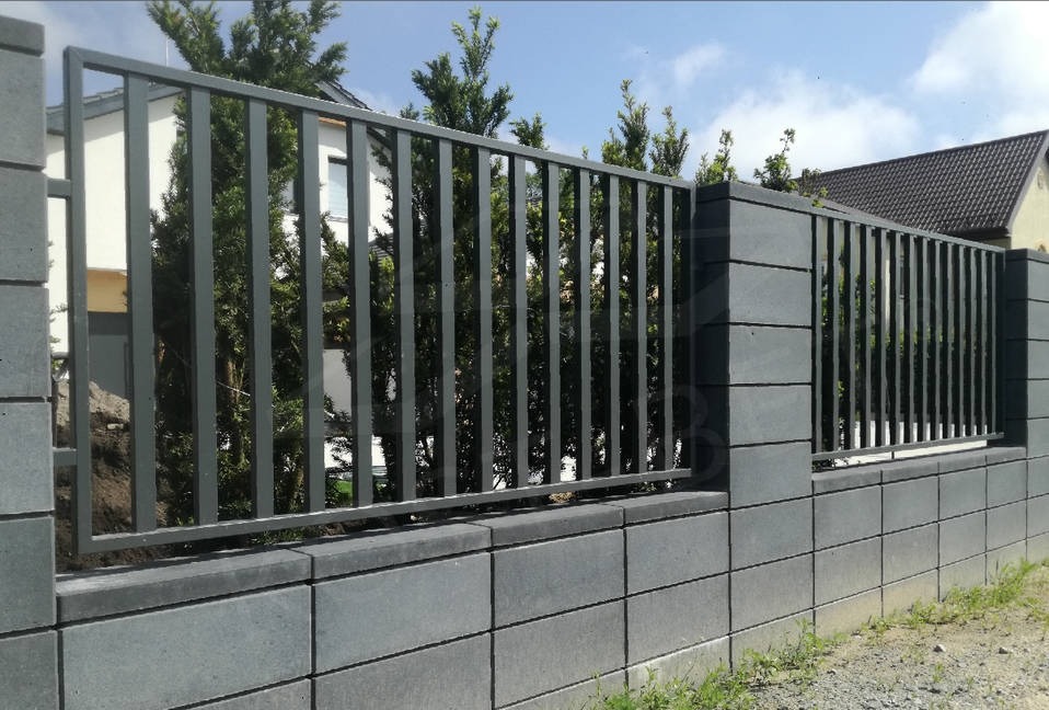 Nowoczesne ogrodzenie metalowe z pionowymi prętami, osadzone na murku z szarej kostki betonowej, widok na dom i drzewa w tle.